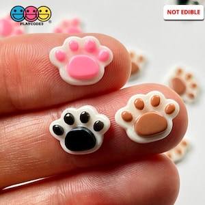 May include: Three small plastic paw print charms in pink, brown, and black. The charms are white with a raised paw print design. The paw prints are approximately 1 inch in diameter. NOT EDIBLE.