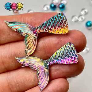 May include: Two iridescent mermaid tail charms with a scale pattern. The charms have a rainbow color scheme with hues of pink, yellow, and blue. The logo "PLAYCODE3" is visible in the top left corner.