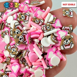 May include: A variety of pink, white, and brown polymer clay charms, including hearts, teddy bears, and circles. The charms are in a hand, and the text "NOT EDIBLE" is visible in the upper right corner of the image.