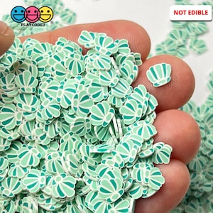 May include: A hand holding a large amount of green and white polymer clay flower shaped sprinkles. The sprinkles are not edible.  NOT EDIBLE is written in red letters on a white background in the upper right corner of the image.