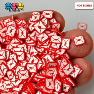 May include: A large pile of red and white envelope-shaped polymer clay charms with a small heart inside each envelope. The charms are not edible. "NOT EDIBLE" is written in white letters on a red background in the upper right corner of the image.