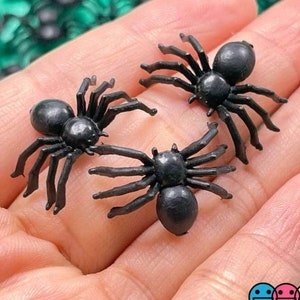 May include: Three black plastic spiders with eight legs and a shiny finish. The spiders are on a hand.