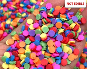 May include: A close-up of a hand holding a large pile of colorful round polymer clay sprinkles. The sprinkles are in various shades of red, orange, yellow, green, blue, and purple. The text "NOT EDIBLE" is visible in the upper right corner of the image.