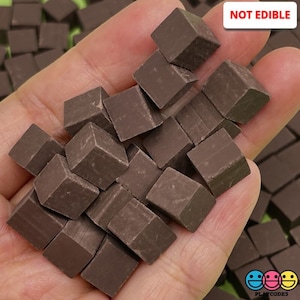 May include: A hand holding a pile of small, dark brown, cube-shaped objects. The cubes are arranged in a random pattern and are all the same size. The text "NOT EDIBLE" is visible in the upper right corner of the image.