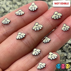 May include: A hand holding a collection of small, yellow bee-shaped charms with black outlines. The charms are not edible. "NOT EDIBLE" is written in black text on a white background in the upper right corner of the image.