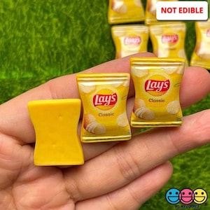 May include: A close-up of a hand holding several miniature yellow and gold Lay's potato chip bags. The bags are labeled "Lay's Classic" and are not edible.