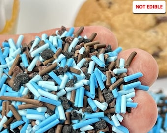 May include: A close-up of a pile of non-edible sprinkles in various shapes and colors. The sprinkles include light blue, dark blue, brown, and black pieces. The text "NOT EDIBLE" is in the top right corner.