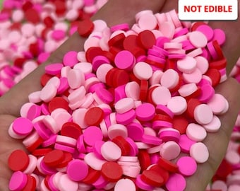 May include: A close-up of a hand holding a large amount of small, round, pink and red sprinkles. The sprinkles are not edible. "NOT EDIBLE" is written in white letters on a black background in the upper right corner of the image.