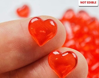 May include: A close-up of a pile of small, red, heart-shaped, clear plastic charms. The text "NOT EDIBLE" is visible in the upper right corner of the image.