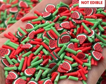 May include: A close-up of a handful of colorful polymer clay pieces. The pieces are shaped like watermelon slices and green sticks. The watermelon slices are red with black seeds and white rinds. The text "NOT EDIBLE" is in the top right corner.