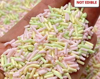 May include: A close-up of a hand holding a pile of pastel pink, yellow, and green sprinkles. The sprinkles are long and thin, and they are arranged in a random pattern. The text "NOT EDIBLE" is visible in the upper right corner of the image.