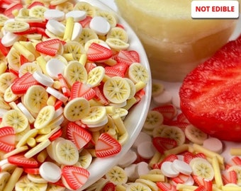 May include: A bowl filled with colorful, decorative, non-edible sprinkles shaped like banana slices, strawberry slices, and white dots. The sprinkles are scattered around the bowl on a white surface. The text "NOT EDIBLE" is visible in the upper right corner of the image.