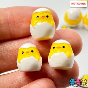 May include: Three yellow chick figurines, each with black eyes and a small orange beak, are peeking out of white eggshells. The figurines are made of plastic and are about 1 inch tall.  NOT EDIBLE.
