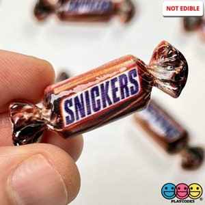 May include: A miniature replica of a Snickers candy bar, wrapped in brown and purple foil with the Snickers logo printed on it. The candy bar is not edible and is labeled "NOT EDIBLE".