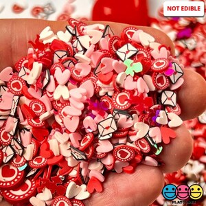 May include: A close-up of a hand holding a variety of red, pink, and white heart-shaped and envelope-shaped polymer clay sprinkles. The sprinkles are decorated with black outlines and red polka dots. The text "NOT EDIBLE" is visible in the upper right corner of the image.