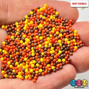 May include: A close-up of a pile of small, round, colorful beads in brown, orange, and yellow. The beads are not edible. "NOT EDIBLE" is written in white text on a black background in the upper right corner of the image.