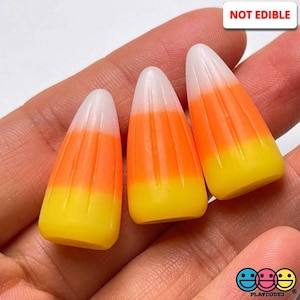 May include: Three candy corn shaped decorations in white, orange, and yellow. The decorations are not edible and have a striped pattern. The text "NOT EDIBLE" is visible in the image.