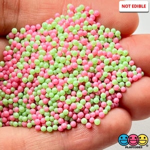 May include: A close-up of a pile of small, round, pink and green beads. The beads are shiny and have a slight shimmer. The text "NOT EDIBLE" is visible in the upper right corner of the image.