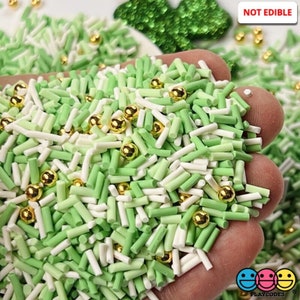 May include: A handful of green, white, and gold sprinkles. The sprinkles are in the shape of small cylinders and spheres. The text "NOT EDIBLE" is in the upper right corner.