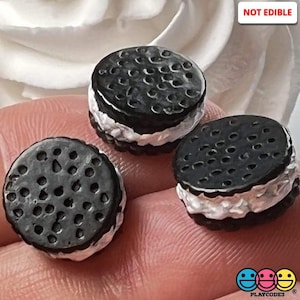 May include: Three miniature chocolate sandwich cookies with white frosting. The cookies are black and have a textured surface. The cookies are not edible. "NOT EDIBLE" is written in white on a black background in the upper right corner of the image.