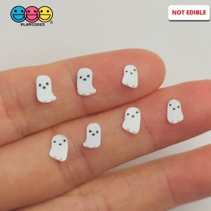 May include: Seven white ghost-shaped charms with black eyes. The charms are small and are being held in a person's hand. The charms are not edible. "NOT EDIBLE" is written in red letters on a white background in the upper right corner of the image.