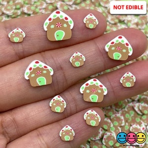May include: A close-up of a hand holding a variety of gingerbread house-shaped charms. The charms are decorated with white icing, red candy dots, and green details. The charms are not edible. The text "NOT EDIBLE" is visible in the upper right corner of the image.
