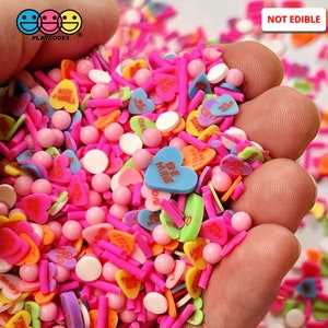 May include: A close-up of a hand holding a variety of colorful heart-shaped sprinkles with messages like "Be Mine", "Hug", "Kiss", and "XOXO". The sprinkles are not edible.