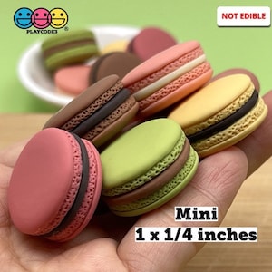 May include: A set of six miniature macarons in various colors, including pink, green, yellow, and brown. Each macaron is approximately 1 inch by 1/4 inch. The macarons are arranged on a hand, with the text "Mini 1 x 1/4 inches" visible below. The text "NOT EDIBLE" is visible in the upper right corner of the image.