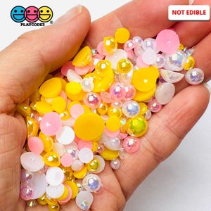 May include: A mix of yellow, pink, and white round and flat resin embellishments. The embellishments are in a hand, and the text "NOT EDIBLE" is in the upper right corner of the image.