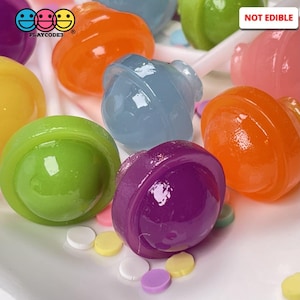 May include: A collection of colorful, round lollipops on a white surface with sprinkles. The lollipops are in various colors, including blue, green, purple, orange, and yellow. The lollipops are not edible and are labeled "NOT EDIBLE".