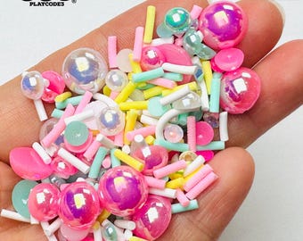 May include: A handful of colorful craft sprinkles and beads in various shapes and sizes. The mix includes iridescent spheres, pink, yellow, and white sticks, and pastel-colored flat pieces. The text "NOT EDIBLE" is visible.