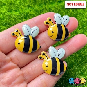 May include: Three yellow and black bee-shaped resin embellishments with white wings. The bees have a smiling face and are perfect for adding a whimsical touch to crafts.