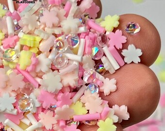 May include: A mix of pastel pink, white, and yellow sprinkles in the shape of flowers and small cylinders. There are also clear iridescent beads. The text "NOT EDIBLE" is visible in the upper right corner of the image.