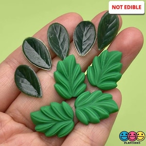 May include: A collection of eight green plastic leaves, some with a smooth surface and others with a textured surface. The leaves are arranged in a hand, with the text "NOT EDIBLE" visible in the top right corner of the image.