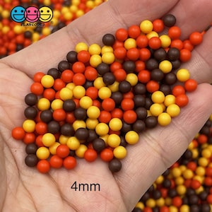 May include: A close-up of a hand holding a pile of 4mm round beads in brown, orange, and yellow colors. The beads are small and uniform in size.