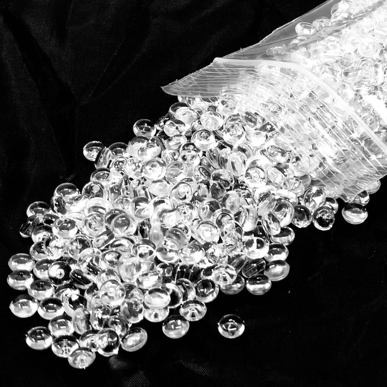 以下が含まれることがあります： 透明なプラスチックバッグから、大量の透明で丸いガラスビーズがこぼれ出ている様子。ビーズは黒い表面に散らばっており、透明なビーズと暗い背景とのコントラストを生み出しています。ビーズは滑らかで反射性があります。