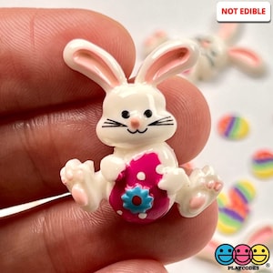 May include: White ceramic Easter bunny figurine holding a pink egg with white polka dots and a blue flower. The bunny has pink ears and a pink nose.