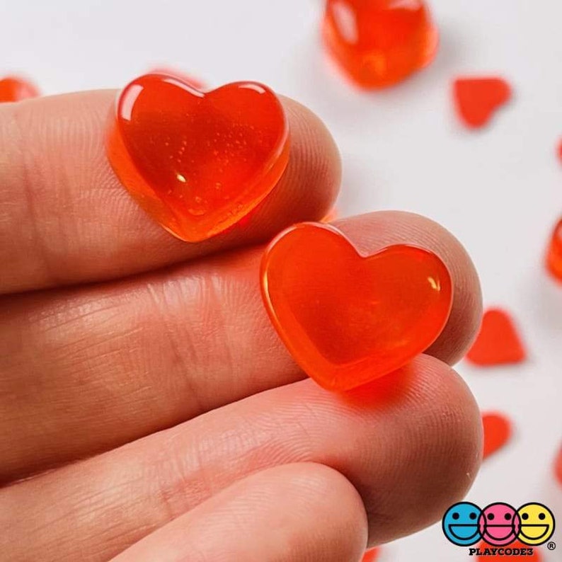 May include: Two bright orange heart-shaped candies, one held in a hand, the other on a white surface.