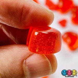 May include: A close-up of a single, bright red, translucent, square-shaped gummy candy. The candy is slightly rounded on the edges and has a shiny, glossy finish.