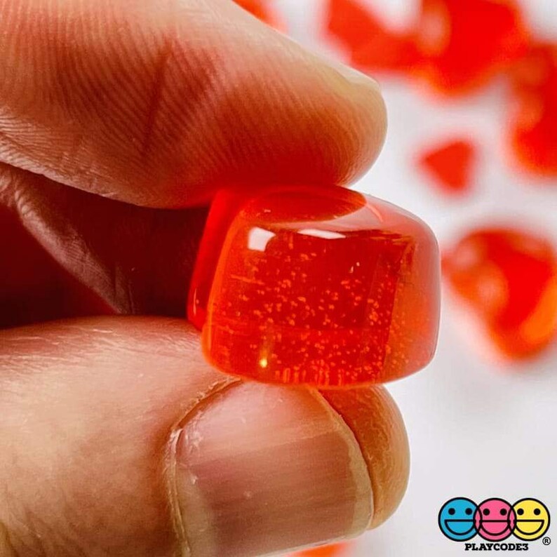May include: A close-up of a single, bright red, translucent, square-shaped gummy candy. The candy is slightly rounded on the edges and has a shiny, glossy finish.
