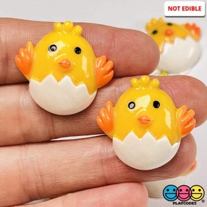 May include: Two yellow chick figurines with orange wings and black eyes, each inside a white cracked egg shell. The figurines are made of resin and are not edible. "NOT EDIBLE" is written in red letters at the top of the image.
