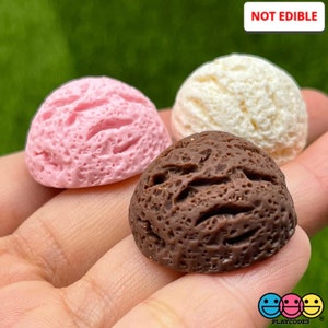 May include: Three miniature ice cream cones in pink, white, and brown. The cones are made of a textured material and are not edible. The text "NOT EDIBLE" is visible in the image.