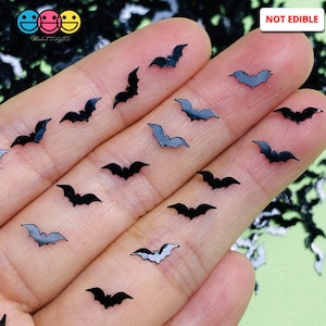 May include: A close-up of a hand holding a variety of black bat-shaped glitter. The glitter is scattered on a green surface. The text "NOT EDIBLE" is visible in the upper right corner of the image.