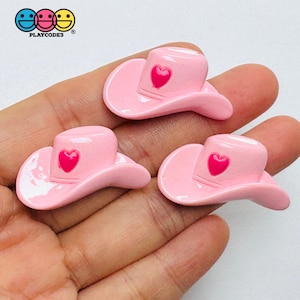May include: Three small, glossy, light pink cowboy hat charms, each featuring a bright hot pink heart emblem centered on the front of the crown. These miniature hat-shaped accessories have a smooth, shiny finish and a curved brim design.