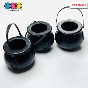 May include: Three small, black cauldron-shaped containers with arched handles. The containers have a textured surface and a circular opening. The top right corner of the image has the text "NOT EDIBLE". The Playcode3 logo is in the top left corner.