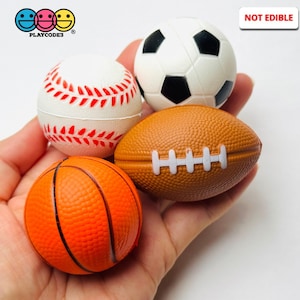 May include: A collection of miniature sports balls, including a baseball, soccer ball, football, and basketball. The baseball is white with red stitching, the soccer ball is white with black pentagons, the football is brown with white stripes, and the basketball is orange with black lines. The text "NOT EDIBLE" is visible.