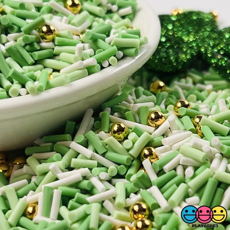 May include: A close-up of a white bowl overflowing with green and white sprinkles and gold metallic beads. The sprinkles are in the shape of small cylinders. A green sequined shamrock is in the background. The image has the Playcode3 logo in the bottom right corner.