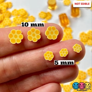 May include: Yellow daisy-shaped resin embellishments in two sizes, 10 mm and 5 mm.  The embellishments are shown on a hand, with the text "NOT EDIBLE" and "PLAYCODES" visible in the background.