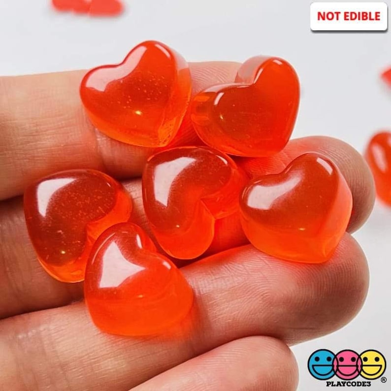 May include: A close-up of a hand holding several small, bright orange heart-shaped plastic charms. The charms are translucent and have a glossy finish. The text "NOT EDIBLE" is visible in the upper right corner of the image.