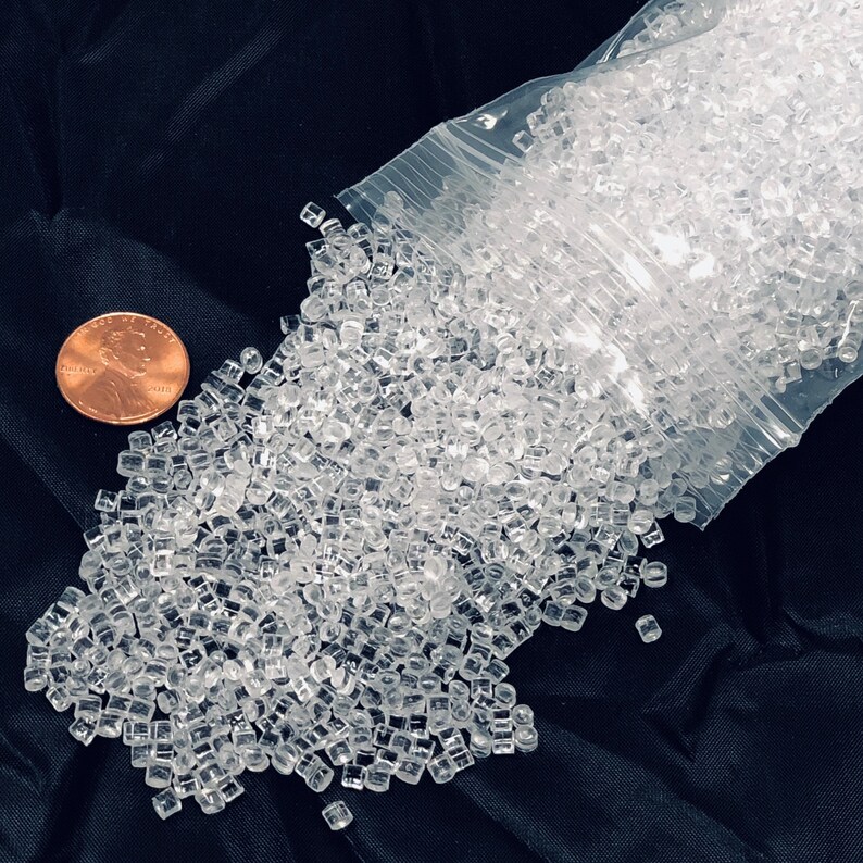 以下が含まれることがあります： 透明な立方体型の小さなプラスチックビーズが、透明なビニール袋からこぼれ落ちています。サイズの比較のために銅貨が隣にあります。ビーズは暗く、質感のある表面に散らばっています。